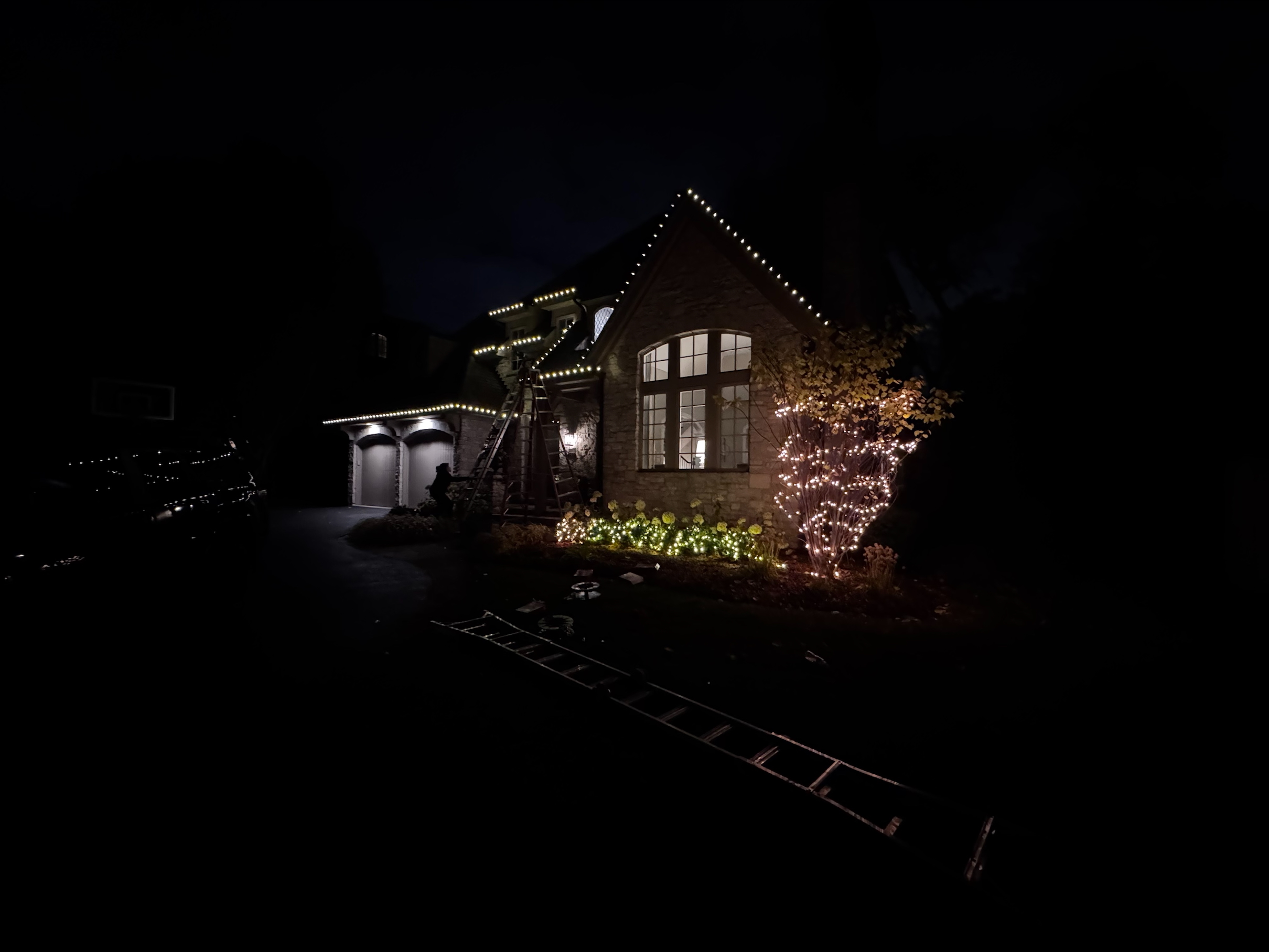Stone home at night with warm white Christmas lights along the roofline, garage, and a wrapped tree