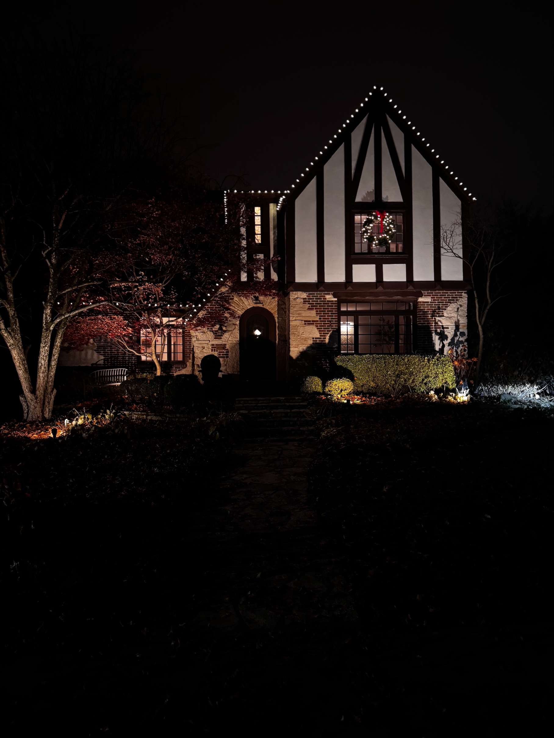 Tudor-style home at night with warm white Christmas lights along the roofline and a lit wreath on the window
